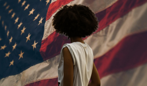 Black woman with natural hair stands in front of a large American flag, symbolizing strength, resilience, and reflection amidst national policy changes